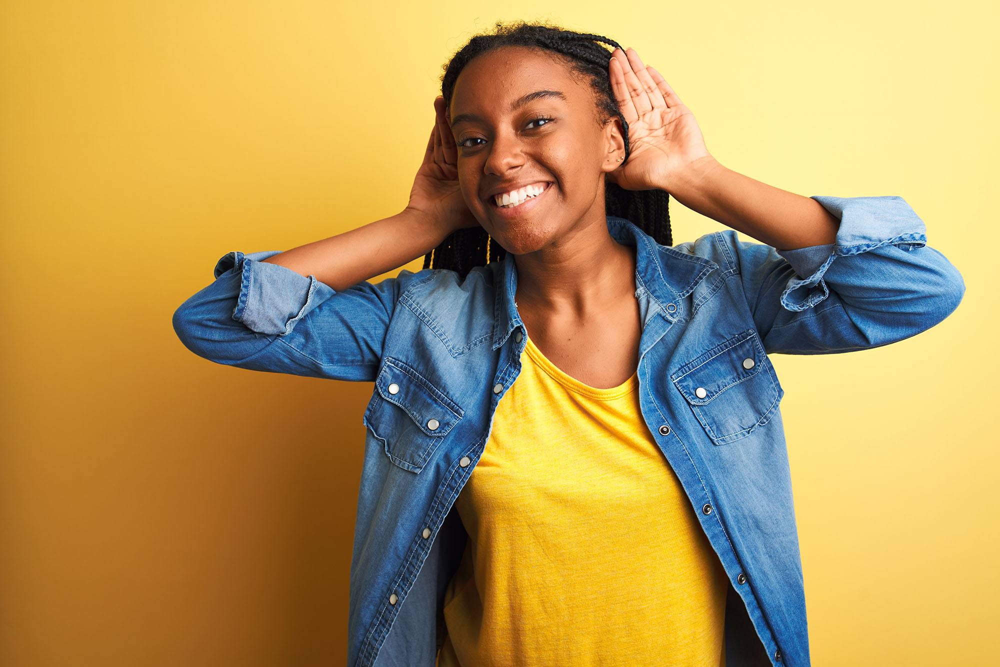 Young african american woman wearing denim shirt standing over isolated yellow background Trying to hear both hands on ear gesture, curious for gossip. Hearing problem, deaf